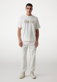 Weißes Baumwoll-T-Shirt mit "GUESS"-Logo in metallic Gold. Kombiniert mit weißen Jogginghosen und beigen Sneakern. Einfaches, lässiges Design.