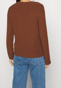 Hnědý pletený svetr s dlouhými rukávy a žebrovanou strukturou, v kombinaci s volnými modrými džíny s klasickým pětidružicovým designem.