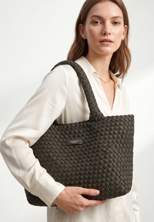 Vrouw met een witte blouse draagt een grote donkere gevlochten draagtas met het label "DIANA&CO FIRENZE" over haar schouder, kijkt neutraal vooruit.