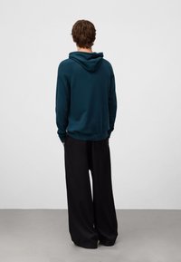Felpa a collo alto teal con coulisse, polsini e orlo a coste. Abbinata a pantaloni neri larghi. Vista da dietro su uno sfondo neutro.