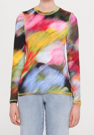 Persoon draagt een nauwsluitend shirt met lange mouwen en een multicolor abstract patroon, gecombineerd met lichtblauwe spijkerbroek, tegen een effen achtergrond.