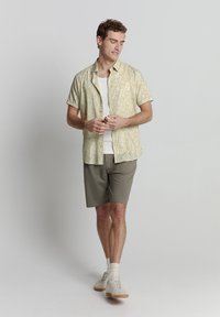 Kurzärmliges, florales Hemd mit Knöpfen in Hellgelb und Grün, kombiniert mit grauen Shorts und weißen Sneakers, bietet eine lockere Passform.