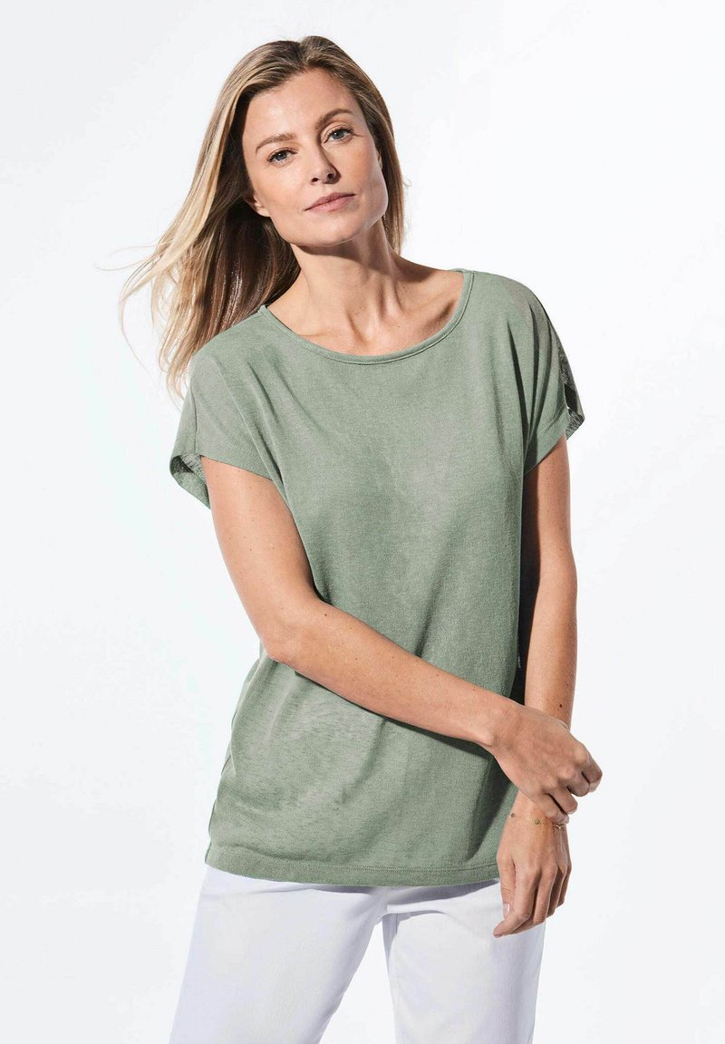 Lässig geschnittenes grünes Jersey-T-Shirt mit Rundhalsausschnitt und kurzen Ärmeln, kombiniert mit weißen Hosen. Glatte Textur mit minimalem Design.