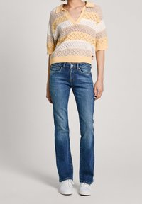 Mujer vestida con una camiseta de polo de punto a rayas en beige, blanco y amarillo, jeans azules y zapatillas blancas, de pie contra un fondo liso.