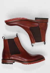 Bottes Chelsea en cuir marron avec panneaux latéraux élastiques, languettes de traction arrière et semelle en caoutchouc rouge à motifs texturés.