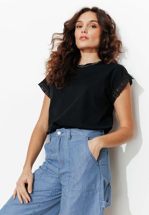 Jeune femme aux cheveux bruns ondulés portant un t-shirt noir avec bordure en dentelle et un pantalon taille haute à rayures bleues et blanches, debout avec une main dans la poche.