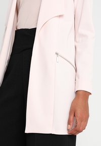 Světle růžový blazer s volným předním dílem, ohnutými rukávy a stříbrným zipem na boku. Vyrobený z hladkého, lehkého materiálu.