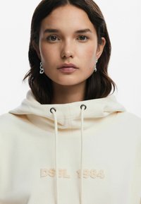 Cream hoodie with drawstrings and black metal eyelets. Features embroidered "DSGL 1984" in matching color, soft fabric texture.