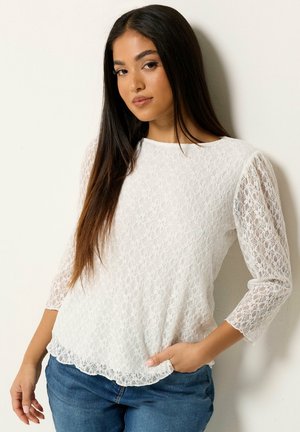 Mujer con cabello largo y oscuro, que lleva una blusa de encaje blanca de manga larga y vaqueros azules, de pie contra una pared clara y lisa.