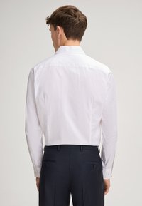 Witte blouse met lange mouwen en een standaardkraag, een gladde textuur en geknoopte manchetten, gecombineerd met een donkere broek. Achteraanzicht.
