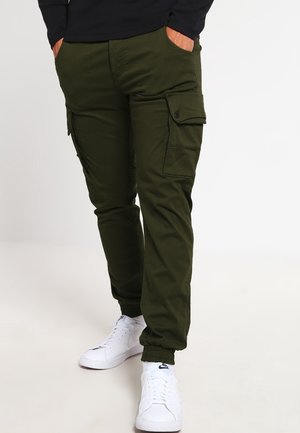 Man wearing olive green cargo pants with elastic cuffs and white Nike sneakers, hands in pockets, standing on a plain light background.