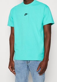 Tyrkysové bavlněné tričko s krátkým rukávem, kulatým výstřihem a černým logem Nike na hrudi. Nosené s modrými džíny, které mají volný střih.