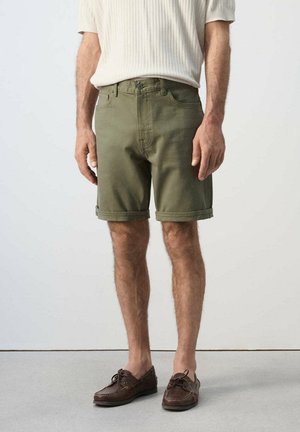 Hombre vestido con pantalones cortos de dobladillo enrollado en verde oliva, camiseta de manga corta de canalé crema y mocasines de cuero marrón, de pie sobre un suelo gris.