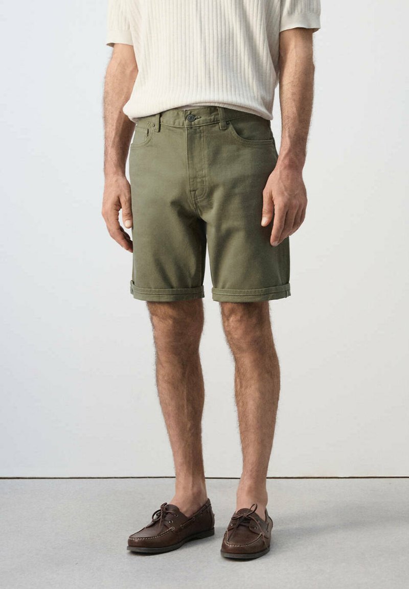 Homme portant un short vert olive à ourlet roulé, une chemise crème côtelée à manches courtes et des mocassins en cuir marron, debout sur un sol gris.