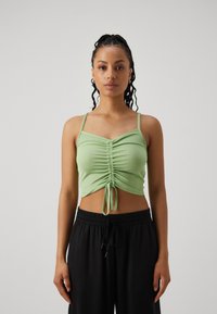 Svetlozelený crop top s nastaviteľným šnúrkou, s riasenouTextúrou a tenkými ramienkami, doplnený o voľné čierne nohavice.