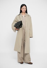 Μπεζ υπερμεγέθη παλτό με μεγάλα πέτα, φορεμένο πάνω από μπεζ ζακέτα με φερμουάρ και αντίστοιχους παντελόνες σε φαρδιά γραμμή. Μαύρη υφαντή τσάντα χειρός και καφέ παπούτσια.