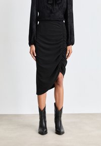 Schwarze knielange Rock mit geraffter Textur auf der Seite und asymmetrischem Saum. Kombiniert mit schwarzen, spitzen Ankle Boots.