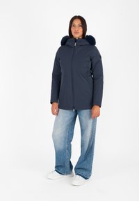 Giacca puffer blu navy con cappuccio bordato in pelliccia, zip frontale e tasche laterali, abbinata a jeans a gamba larga in denim chiaro e scarpe da ginnastica bianche.