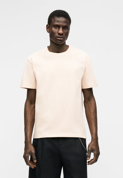 Άνδρας που φοράει απλό, ανοιχτό μπεζ κοντομάνικο μπλουζάκι και μαύρο παντελόνι, σε απλό ανοιχτό γκρι φόντο.