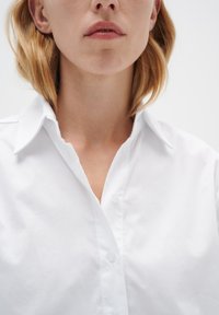 Camisa blanca de botones con cuello clásico, presenta una textura de algodón suave y cierre frontal con botones, vista desde el pecho hacia arriba.