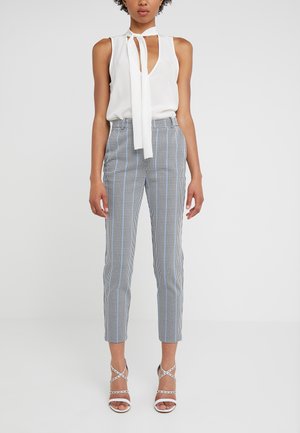 Femme portant un chemisier blanc sans manches avec un nœud au cou, un pantalon slim court à motifs et des sandales blanches à talons hauts avec brides, debout devant un fond uni.