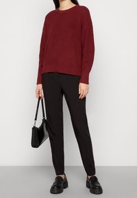 Maroon ribbed knit sweater with long sleeves, paired with black tapered trousers and chunky black loafers. Model holds a black handbag.