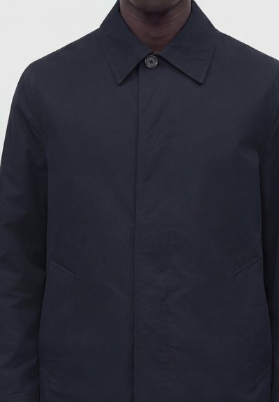 Gros plan d'une personne portant un manteau bleu marine foncé avec un col et un seul bouton visible fermé au niveau du cou.