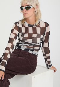 Personne aux cheveux blonds clairs et lunettes de soleil, portant un haut transparent à manches longues avec un motif géométrique marron et blanc et un pantalon en velours côtelé marron, assise sur un bloc blanc.