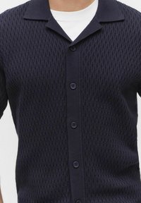 Marineblauwe gebreide cardigan met een ruitpatroon, korte kraag en knoopsluiting. Heeft vijf donkere knopen langs de voorkant en een gestructureerde stof.