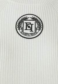 Maglione bianco a costine con un patch circolare ricamato nero che presenta un logo a doppia 'E' e accenti di alloro.