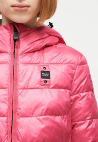 Γυναίκα που φοράει ένα φωτεινό ροζ μπουφάν με κουκούλα, μερικώς φερμουαρ, με μια μικρή μαύρη ετικέτα «Blauer U.S.A.» στο στήθος.