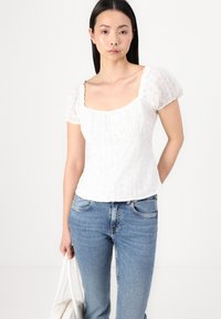 Blouse en coton blanc avec des manches bouffantes, décolleté carrée et détails de broderie œillet. Portée avec un jean bleu, accompagnée d'un sac à main blanc.