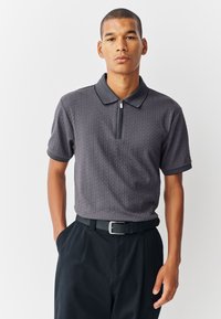 Grijs poloshirt met korte mouwen en een ritskraag, voorzien van een gestructureerd patroon. Gecombineerd met een zwarte broek en een zwarte riem.