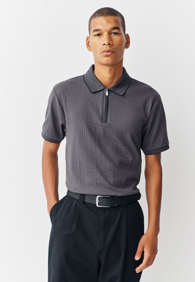 Grijs poloshirt met korte mouwen en een ritskraag, voorzien van een gestructureerd patroon. Gecombineerd met een zwarte broek en een zwarte riem.