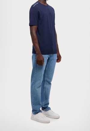 Camiseta azul marino de manga corta, con cuello redondo; en la parte frontal presenta un logotipo sutil; combinada con jeans azul claro y zapatillas blancas.