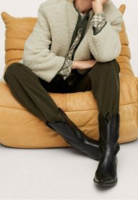 Personne portant des bottes de cowboy en cuir noir, un pantalon vert olive, une chemise à carreaux et une veste sherpa beige clair assise sur une chaise rembourrée beige.