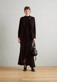 Μαύρο σακάκι συνδυασμένο με εφαρμοστό μαύρο φόρεμα που διαθέτει φαρδύ τελείωμα, φορεμένο με μαύρες μποτές μέχρι τον αστράγαλο και μικρή τσάντα χειρός. Λείες υφές.