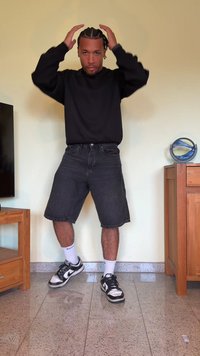 Sweatshirt preto, calções de ganga escuros, meias brancas, sapatilhas pretas e prateadas. Cabelo estilizado em tranças. Em pé numa sala com móveis de madeira.