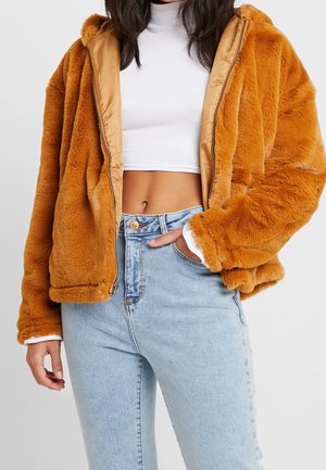 Vrouw die een cropped witte coltrui draagt, lichtblauwe high-waisted jeans en een oranje nepbontjas met capuchon die open is.