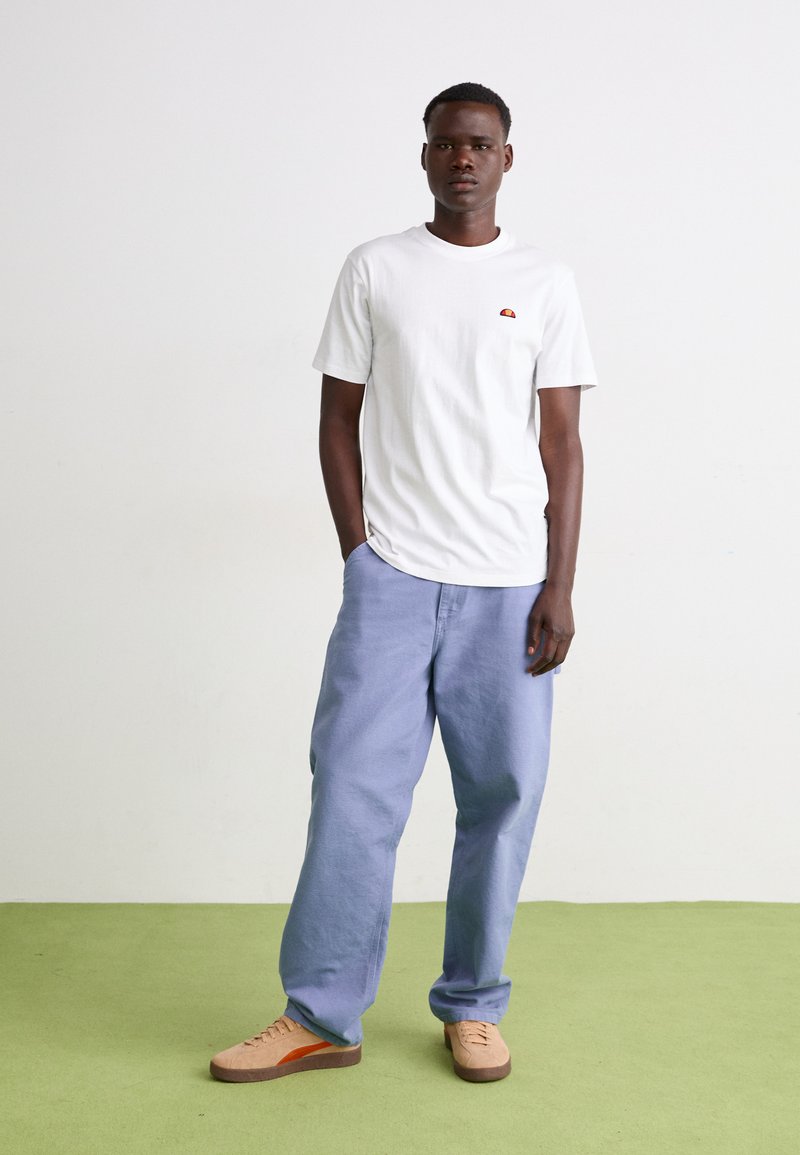 Biała bawełniana koszulka z małym pomarańczowym logo, w połączeniu z luźnymi jasnoniebieskimi spodniami i beżowymi butami z brązowymi podeszwami. Tło w kolorze zielonym.