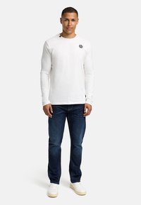 Weißes langärmliges T-Shirt mit schwarzem Logo auf der Brust, kombiniert mit dunkelblauen Jeans und weißen Sneakers. Einfaches, lässiges Design.