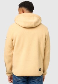 Sudadera de felpa beige con una capucha grande y tejido texturizado. Presenta una etiqueta tejida en el dobladillo; combinada con jeans de mezclilla azules.