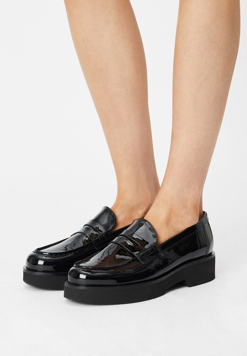 Mocassins en cuir verni noir avec une semelle plateforme surélevée, présentant un détail cousu et une finition lisse.