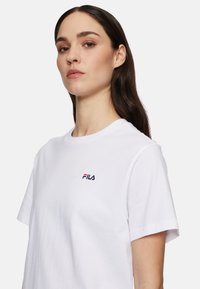 T-shirt bianca in cotone con scollo rotondo. Presenta un piccolo logo Fila in blu navy e rosso sul lato sinistro del petto. Maniche corte, texture liscia.