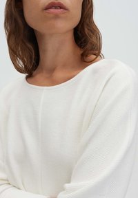 Vrouw met golvend bruin haar, gekleed in een geribd wit topje met lange mouwen, gefotografeerd van schouders tot lippen tegen een effen lichte achtergrond.