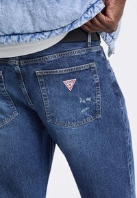 Jeans in denim blu scuro con un logo triangolare rosso e bianco "GUESS?" sulla tasca posteriore, con texture visibile e leggero sbiadimento.