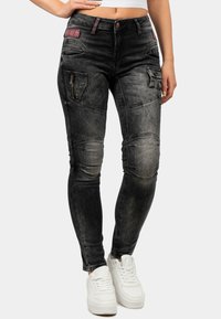 Jeans in denim nero con finitura sbiadita, dotati di tasche stile cargo, dettagli sulle ginocchia e una tasca con cerniera. Abbinati a sneaker bianche.