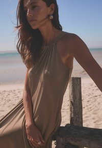 Robes halter en beige avec un détail de décolleté froncé, en tissu doux et silhouette fluide, accompagnées de boucles d'oreilles en or et d'un arrière-plan de plage.