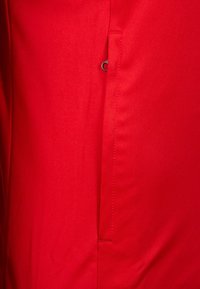 Rode stof, gladde textuur, strakke ontwerp met een zijzak met een metalen klipsluiting. Strakke lijnen met een minimale afwerking.