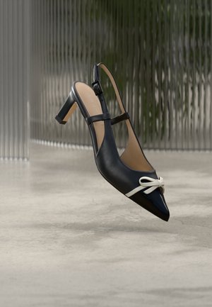 Chaussure à talon haut noire avec bride arrière, bout pointu et détail nœud blanc, présentée sur un fond gris texturé.